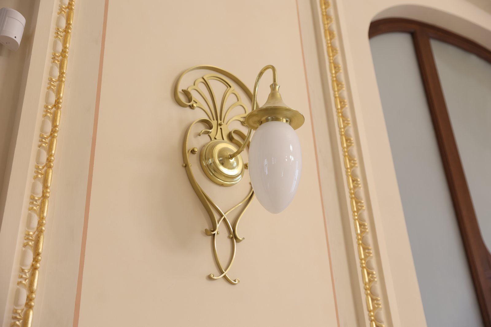 An Art Nouveau brass wall sconce with an opal glass globe, on a white wall with gilded mouldings