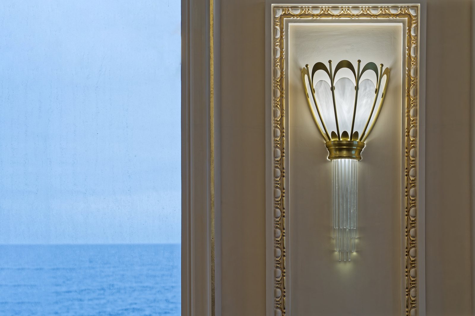A lit Art Nouveau sconce on a wall with gilded mouldings, beside a window looking out to sea