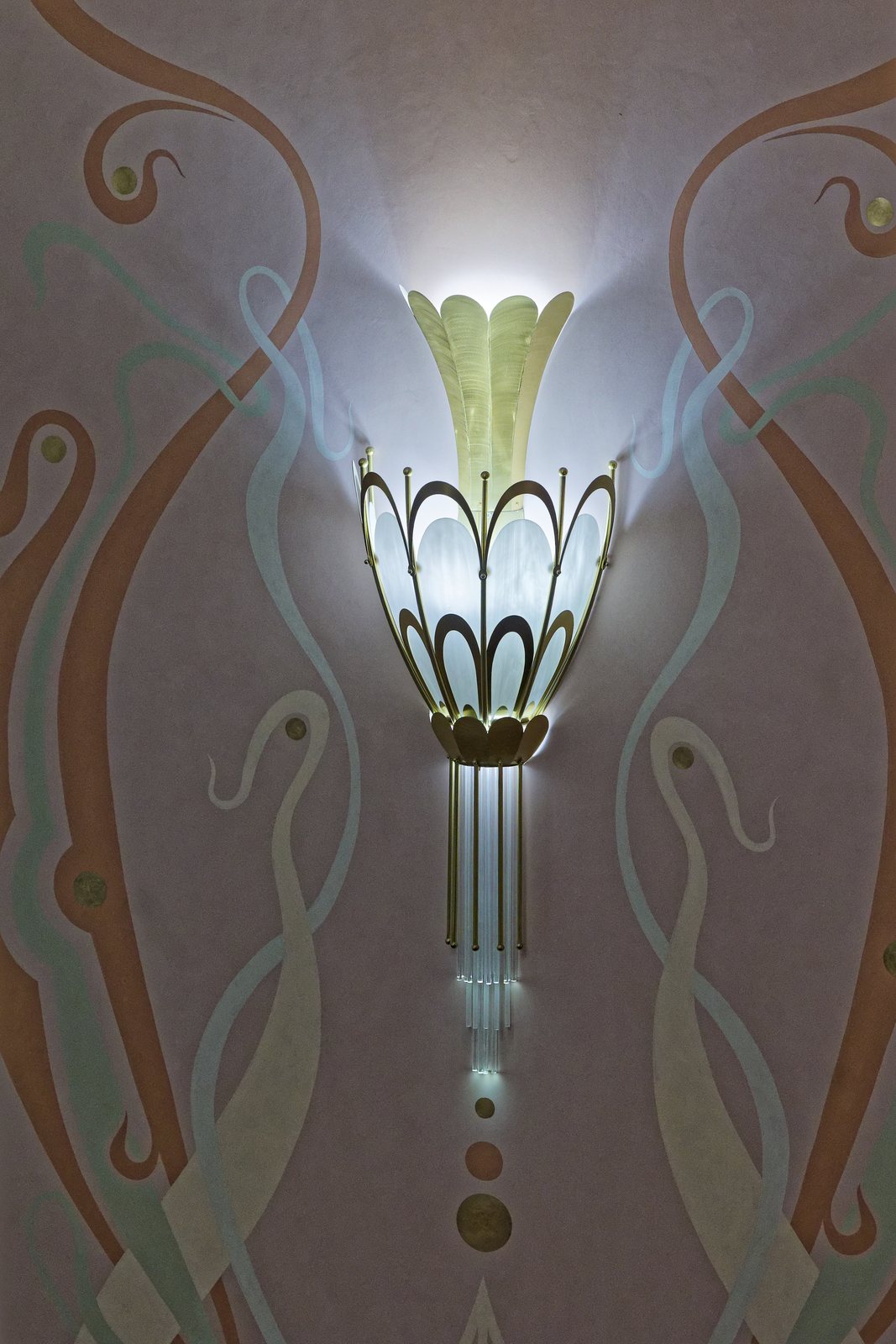 A lit Art Nouveau wall sconce, on a wall with painted decoration in pastel tones