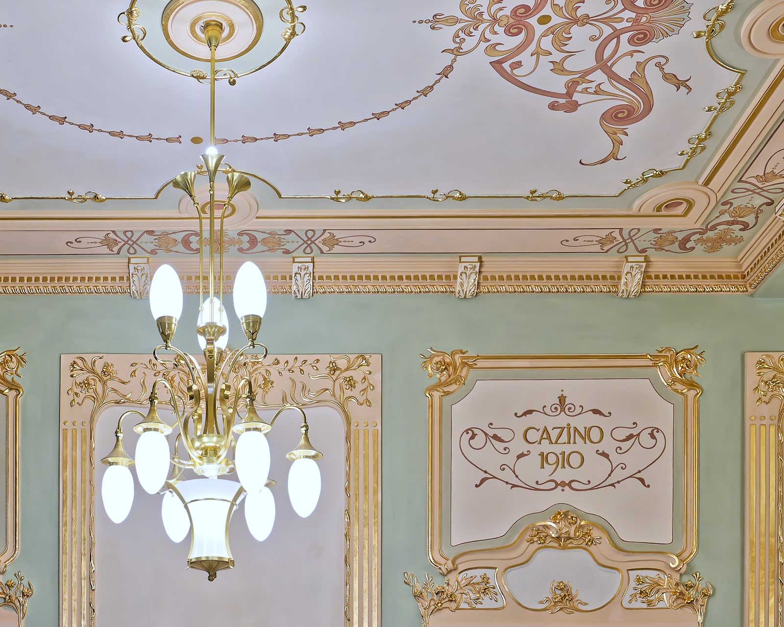 A brass chandelier with arms and opal glass globes, beneath the painted ceiling bearing the inscription “Cazino 1910”