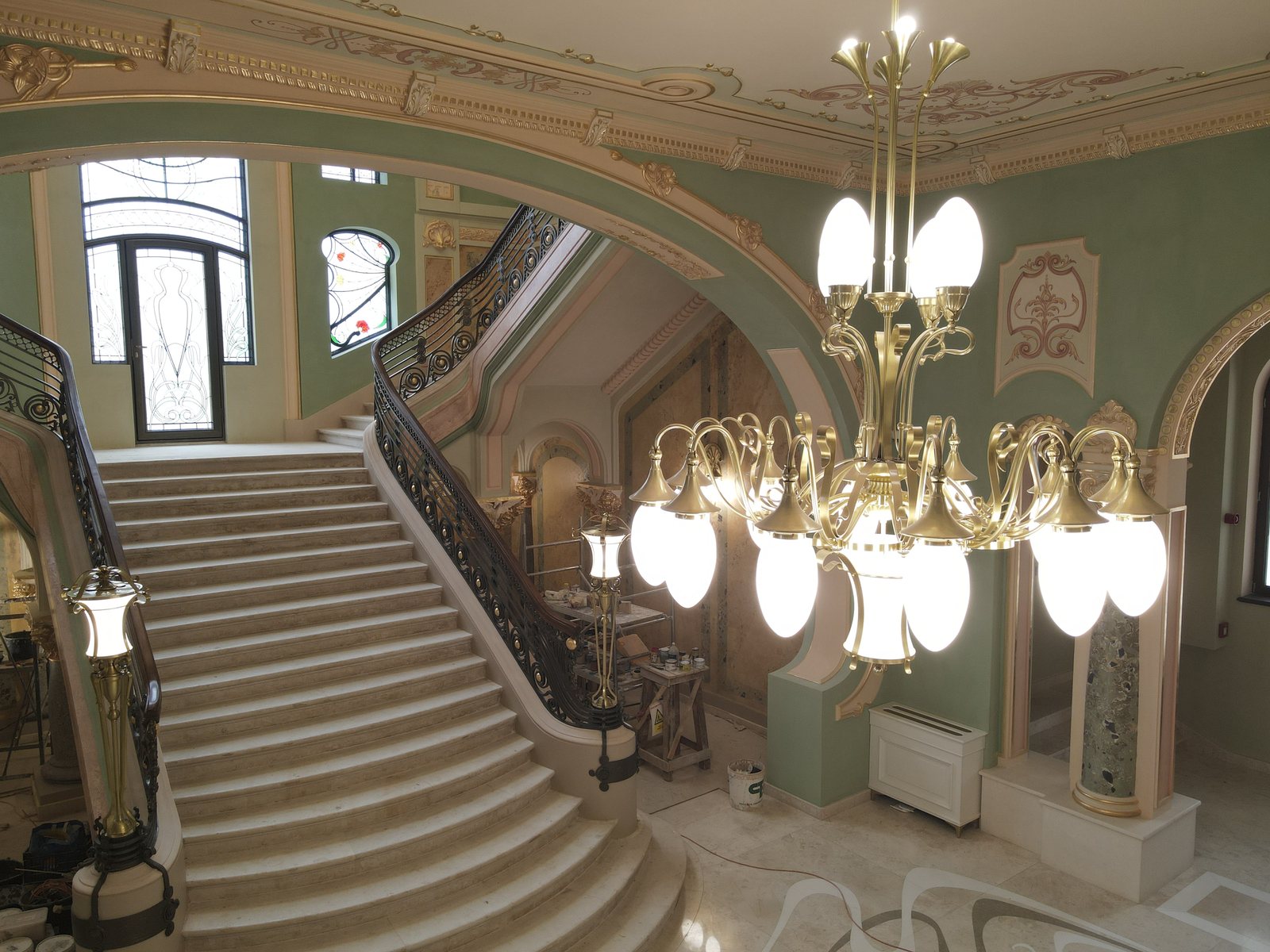 A brass chandelier with arms and glass globes, photographed at its own height, with a marble staircase behind