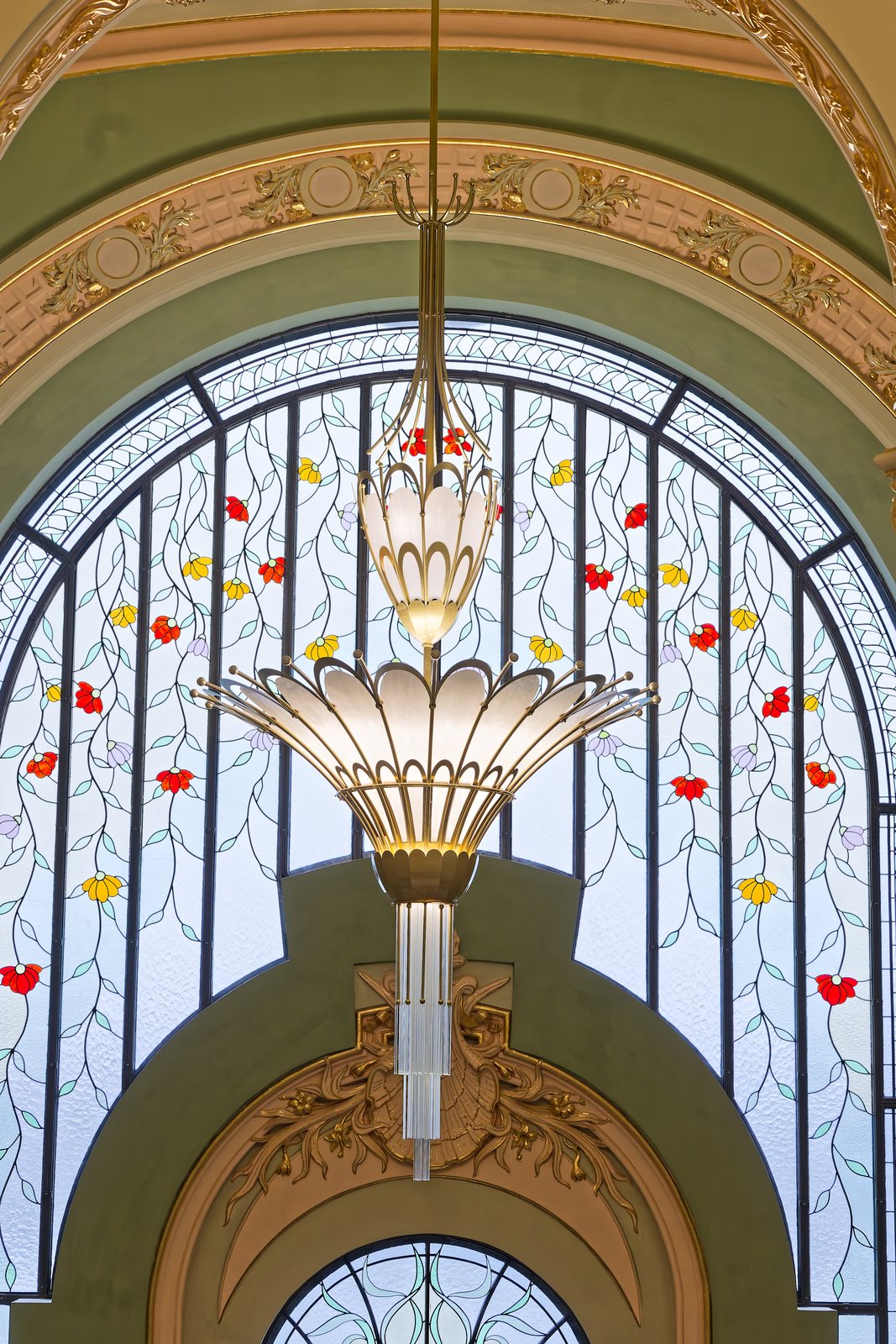 An Art Nouveau chandelier hanging in front of a large stained-glass window with floral motifs, on the grand staircase