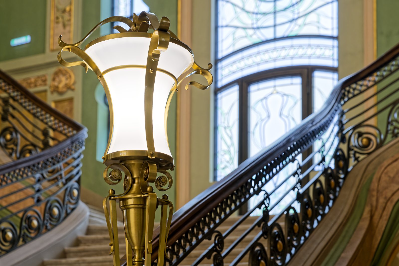 A brass floor lamp with an opal glass globe at the foot of the grand staircase, the stained glass behind it