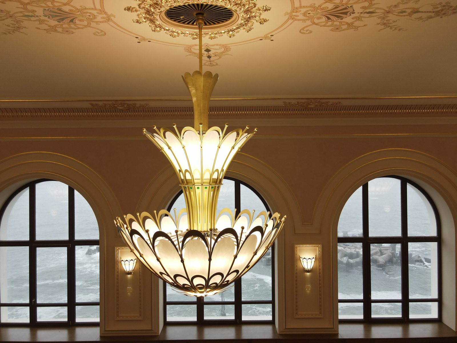 An Art Nouveau chandelier in brass and opal glass, photographed at its own height, with the sea visible through the arched windows