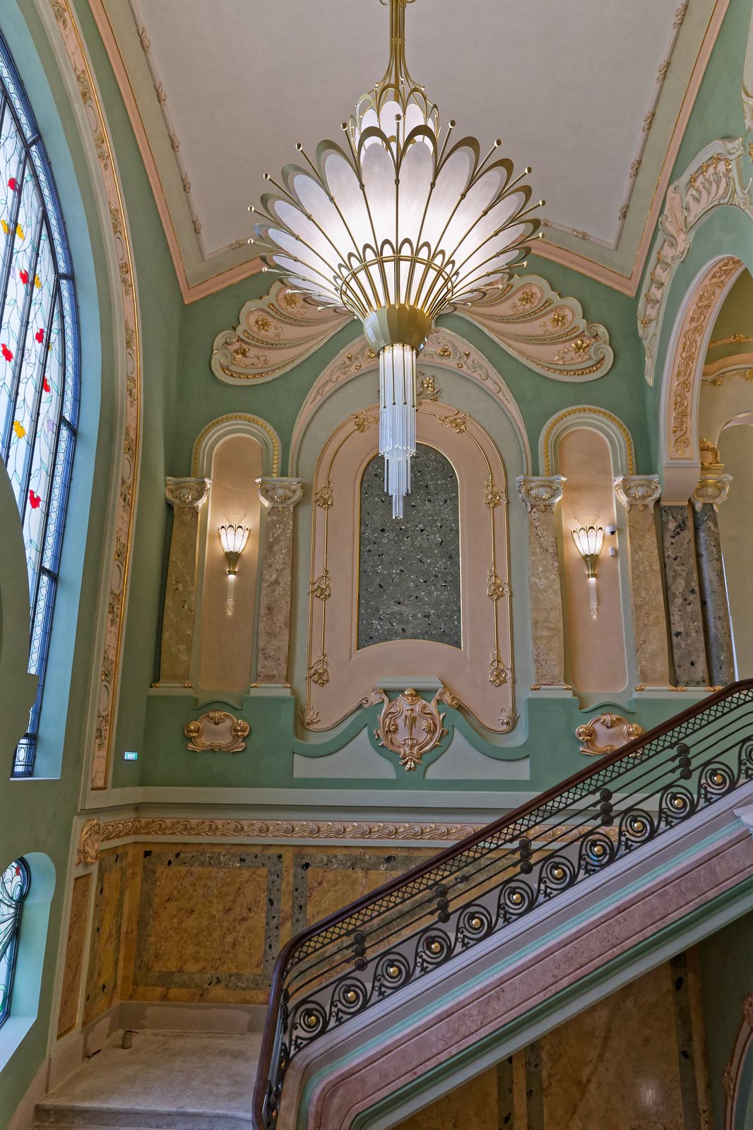 An Art Nouveau chandelier and lit sconces on the grand staircase, with the wrought-iron balustrade in the foreground