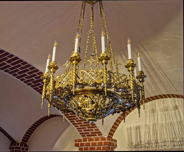 A brass chandelier with 12 arms, under the white vault with exposed brick ribs of the narthex