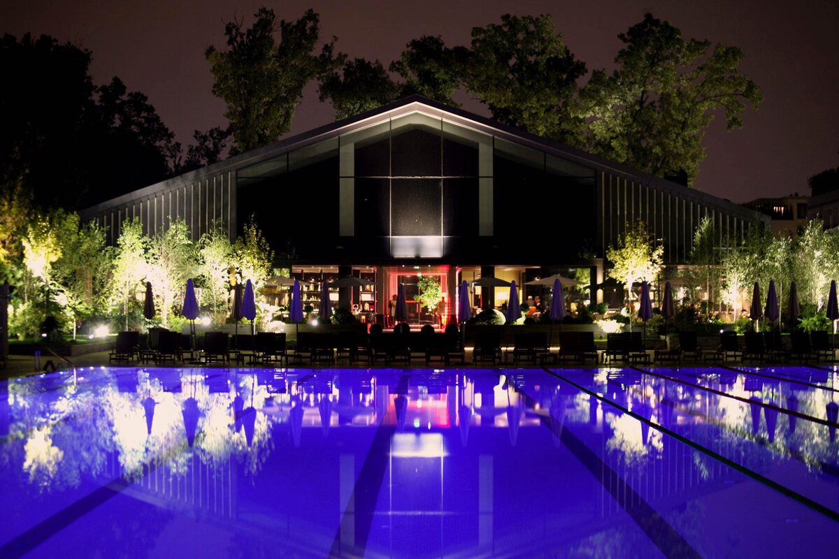 The outdoor pool at Stejarii Country Club at night, with the lit restaurant pavilion behind it and the trees of the Baneasa forest all around